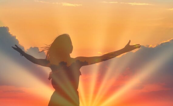 Individual standing in the sunrise with arms lifted toward the sun during summer solstice ritual.